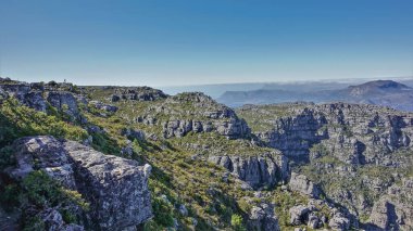 Masa Dağı 'nın tepesinde küçük bir adam silueti görülüyor. Çatlak gri taşların üzerinde az miktarda bitki örtüsü var. Mavi gökyüzü. Cape Town 'da. Güney Afrika