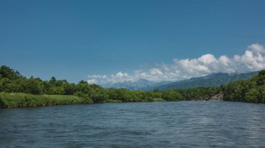 Mavi nehir huzur içinde akar. Uzakta, insanlarla dolu şişme botlar. Bereketli bitki örtüsünün kıyılarında, kır çiçekleri. Gökyüzü ve bulutların arkasındaki resim gibi dağlar. Kamçatka.