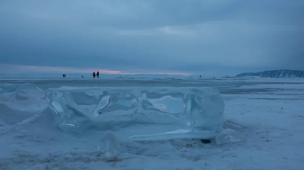 一个闪闪发光的浮冰浮在一个结冰覆盖的湖面上 在远处 在阴云密布的夜空的背景下 可以看到人的微小轮廓 贝加尔湖 — 图库照片