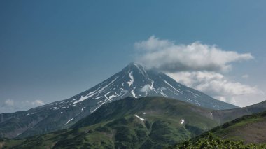 Mavi arka planda güzel bir konik volkan. Yokuşta kar var. Zirvedeki bulutlar. Önde yeşil tepeler var. Kamçatka. Stratovolcano Vilyuchinsky.