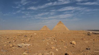 Giza 'nın iki büyük piramidi, bulutlu mavi gökyüzünün arka planına karşı çölde yükselir. Şehirdeki evlerin siluetleri ufukta görünüyor. Ön planda kumlu kayalık toprak var. Kahire. Mısır
