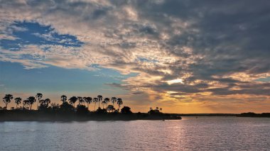 Zambezi nehrinin üzerinde parlak gün batımı. Sakin suyun yüzeyinde güneşin yansımaları, bulutların gölgeleri vardır. Mavi gökyüzüne karşı palmiye ağaçlarının siluetleri. Zimbabwe.