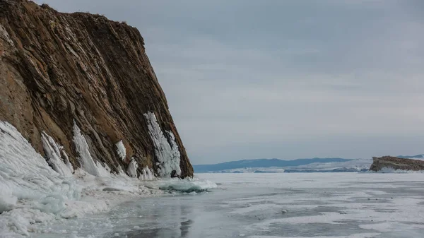 一座没有植被的花岗岩岩石耸立在一个结冰的湖上 底部是冰的 多云的天空映衬下的山脉 冰上的倒影贝加尔湖 — 图库照片