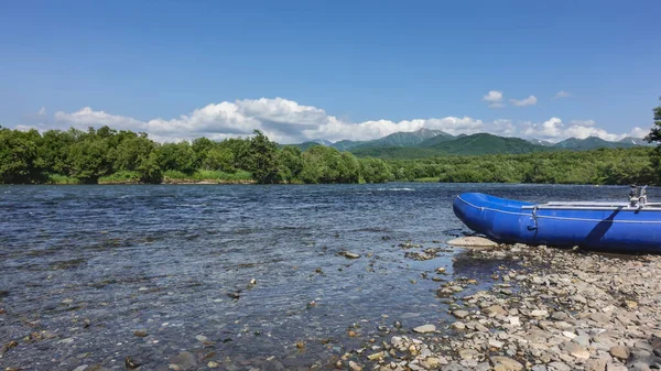 The river flows along a rocky bed. There is an inflatable boat for rafting on the shore. Lush green vegetation in the distance. A picturesque mountain range against the blue sky. Kamchatka