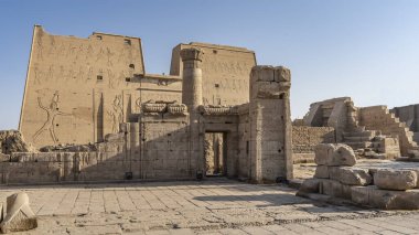 Edfu 'daki Horus Tapınağı. Mavi gökyüzünün arka planında, oymalı yüksek duvarlar, bir sütun, antik taşlar görülebilir. Mısır