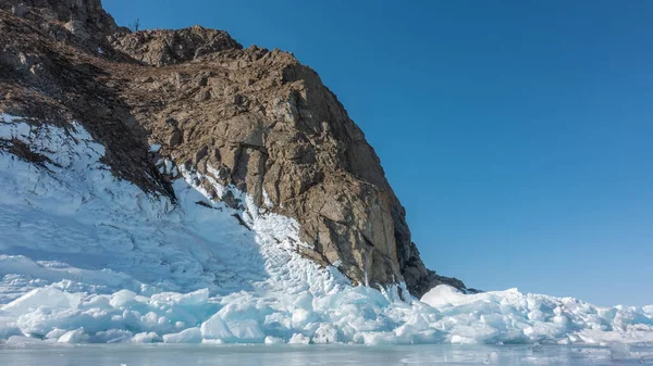 在蓝天的映衬下 一块没有植被的风景如画的花岗岩岩石 石坡上有裂缝 在底部 在一个结冰的湖上 冰块散落着 贝加尔湖 — 图库照片