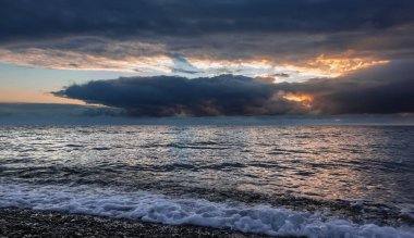 Gökyüzü neredeyse tamamen koyu yoğun bulutlarla kaplıdır. Molalarında güneş ışınlarından parlak turuncu bir ışık var. Denizin parlak yüzeyinde yansıma. Uzakta yağmur yağıyor..