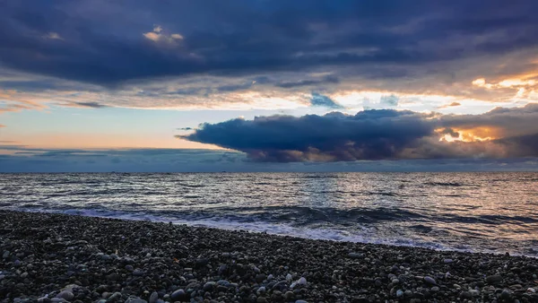 İyi akşamlar. Batan güneşin ışınları yoğun mavi bulutların ardından parlıyor. Gökyüzü turuncudur. Denizin parlak yüzeyinde yansıma. Plaj taşlarında köpük dalgaları. Gün batımı. Karadeniz.