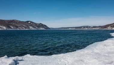 Kar, dondurucu olmayan Angara nehrinin kıyısında uzanıyor. Mavi suda dalgalanmalar var. Karla kaplı dağlar uzaktan görülebilir. Gök mavisi. Rusya