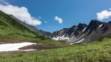Mavi gökyüzüne karşı Vachkazhets dağlarının resimli sirki. Yamaçlarda kar olan yerler var. Alp çayırında yeşil çimenler, kır çiçekleri var. Güneşli bir yaz günü. Kamçatka.