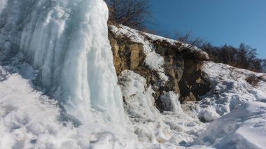 Tepenin yamacında eriyen kar ve buz sarkıtları. Kayalıklardaki kuru çimenler. Güneşli bir gün. Mavi gökyüzü. Baharın başında. Sibirya.