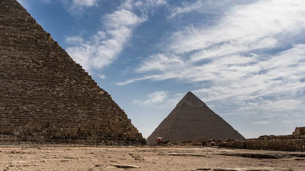 在蓝天和云彩的背景上 两座巨大的吉萨金字塔 切普斯金字塔和切弗伦金字塔 墙壁上的石墙是可见的 脚上有马套在推车上 — 图库照片