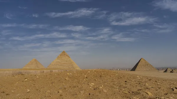 ギザの3つの大きなピラミッド 青い空と雲の背景を背景に砂漠の砂の上にCheops Chephren Mikerinが上昇します 水平線には近代カイロの建物が見える エジプト — ストック写真