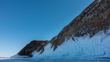 Granit kaya, bitkilerden yoksun, mavi gökyüzüne karşı. Taban buz saçakları ve sıçramalarla kaplı. Donmuş bir gölün yüzeyindeki buz parçaları. Baykal.