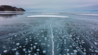 Çatlaklar ve donmuş metan gazı kabarcıkları donmuş gölün pürüzsüz şeffaf buzunda görülebilir. Uzakta, bulutlu bir gökyüzüne karşı bir dağ sırası var. Baykal.