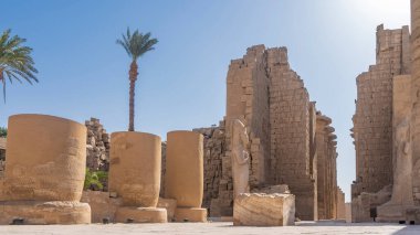 Luxor 'daki antik Karnak Tapınağı. Taş duvarlar ve mavi gökyüzüne karşı sütunlar arasındaki geçit. Kenarlarında firavun heykelleri var. Ön planda, harap olmuş sütunlar. Mısır
