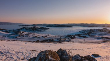 Sibirya 'da kış sabahı. Gökyüzü turuncuyla işaretlenmiş. Donmuş bir gölün üzerindeki güneşin parlaklığı. Yerde kar var. Kıyıya yakın bir turist üssünde evler var. Baykal.