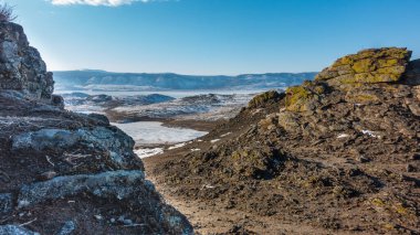 Donmuş göl dağlarla çevrilidir. Tepelerde kar var. Granit kayalar ön planda. Kırık taşların üzerindeki sarı zambaklar. Mavi gökyüzü. Baykal..