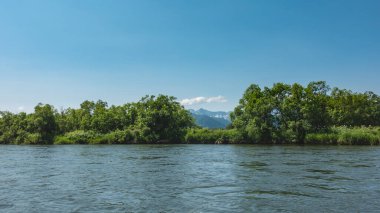 Mavi nehir sakince akar. Sudaki dalgalanmalar. Kıyıda yemyeşil bitki örtüsü var. Gök mavisi gökyüzüne karşı dağlar. Kamçatka..