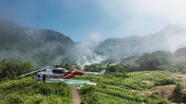 The helicopter is on the landing pad in the Valley of Geysers. There is a meadow with green grass and wildflowers around. Steam from hot springs rises from the gorge above the mountains. Kamchatka