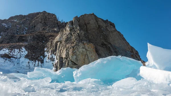 花岗岩在蓝天的映衬下结成岩石 陡坡上的裂缝和雪 在脚下一个结冰的湖上 有一块大冰块 一个阳光灿烂的冬日 贝加尔湖 — 图库照片