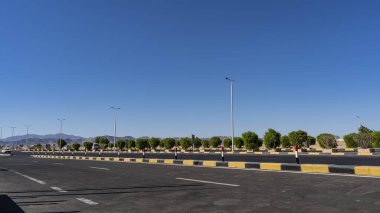 Asfalt yolda beyaz işaretli çizgiler var. Ortada siyah ve sarı bir ayrılma bariyeri var. Yolun kenarındaki yeşil çalılar var. Uzakta, mavi gökyüzüne karşı - dağlar. Mısır