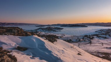 Sibirya 'da kış sabahı. Gökyüzü turuncuyla işaretlenmiş. Donmuş bir gölde güneş parlıyor. Ön planda parlak karla kaplı kayalık bir tepe var. Baykal. Altın Saat