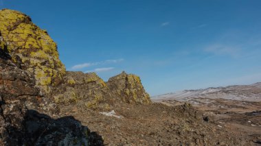 Mavi gökyüzüne karşı granit kayalar. Yamaçlarda çatlaklar ve sarı zambaklar. Uzakta karla kaplı bir dağ sırası. Güneşli bir kış günü. Uzayı kopyala Sibirya 'da. Olkhon.