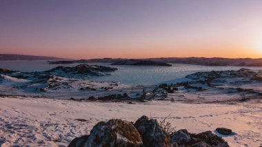 Kış Sibirya şafağı. Gökyüzü pembemsi. Donmuş bir gölün buzunda güneş parlıyor. Sahildeki kamp alanında. Yerde kar ve kuru çimenler var. Önünde kertenkele olan bir kaya. Baykal.