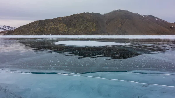 冰封的湖面光滑明亮 冰上可见裂缝和甲烷气泡 水面上的小雪 海岸山脉与多云的天空相映衬 贝加尔湖 — 图库照片