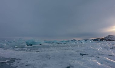 Turquoise ice hummocks are scattered in disorder on the surface of the frozen lake. The glare of the setting sun on the edges. A mountain range against the background of an evening cloudy sky. Baikal