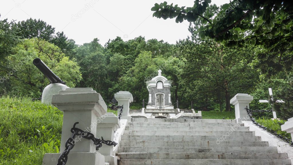 An old chapel of gray-white stones stands in a park on a hill. Around- thickets of green trees. A ladder with a chain railing goes up. Petropavlovsk-Kamchatsky