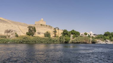 There is green vegetation on the banks of the Nile. An old mausoleum is visible on a sand dune. Clear azure sky. Reflection on the blue water. Egypt. Aswan