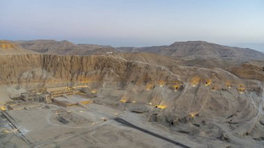 Aerial view of the funerary temple of Pharaoh's Queen Hatshepsut. The ancient building is surrounded by steep cliffs. Electric lighting. Blue dawn sky. Luxor. Egypt