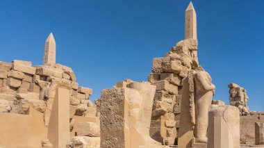 A fragment of the ancient Karnak temple in Luxor. Dilapidated stone walls and sculptures are visible. High obelisks against the blue sky. Egypt