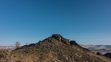 A rocky hill against a clear blue sky. A snow-covered mountain range in the distance. Dry grass and bare trees in the foreground. Siberia. Copy Space