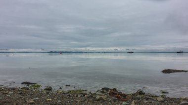 Pebbles are scattered on the shore of the Pacific Ocean. Ships are visible in the distance. The mountain range is hidden in the clouds. Reflection on smooth water. Petropavlovsk-Kamchatsky