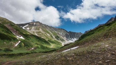 A picturesque mountain range against a background of blue sky and clouds. Patches of snow and green grass on the slopes. Kamchatka. Vachkazhets 