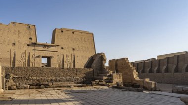 Edfu 'daki Horus Tapınağı mavi gökyüzüne karşı. Yüksek duvarda tanrıların devasa oymalı görüntüleri görülebilir. Harabeler ön planda. Mısır