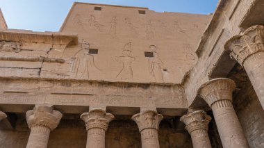 Edfu 'daki Horus tapınağının bir parçası. Çeşitli şekillerde büyük harflerle sütunlar görünür. Yüksek duvarda tanrıların oymaları var. Mavi gökyüzü. Mısır