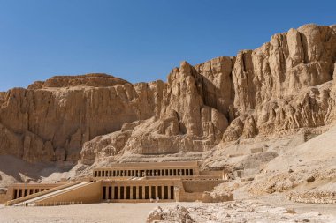 Firavun 'un kraliçesi Hatshepsut' un eski cenaze tapınağı. Merdivenler, sütunlar, heykeller görünür. Mavi gökyüzüne karşı sarp, resimli bir kaya. Mısır