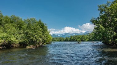 Mavi nehir yemyeşil bitki örtüsüyle bankalar arasında akar. Gökyüzü ve bulutların arkaplanına karşı pitoresk bir sıradağ. Kamçatka.