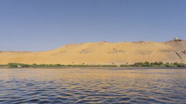 Nil kıyısında dar bir yeşil bitki örtüsü şeridi görünür. Mavi gökyüzüne karşı yüksek bir kum tepesi. Sudaki dalgalar ve röfleler. Mısır