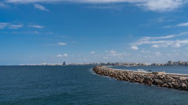 Alexandria 'da deniz manzarası. Akdeniz 'de taş bir iskelede yatlar görülebilir. Uzakta, ufukta, şehir binalarıyla dolu bir set var. Bulutlu mavi gökyüzü. Mısır