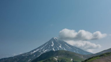 Mavi gökyüzüne karşı karla kaplı yamaçları olan pitoresk bir konik volkan. Bulutlar tepede asılı. Öndeki yeşil tepeler. Uzayı kopyala Kamçatka. Stratovolcano Vilyuchinsky