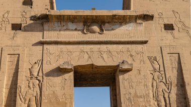Edfu 'daki antik Mısır tapınağı Horus' un bir parçası. Taş duvarlarda tanrıların ve hiyerogliflerin oyulmuş görüntüleri görülebilir. Kapıda mavi gökyüzü var. Mısır
