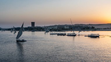 Nil 'de günbatımı. Güneş bir kum tepesinin arkasına saklandı. Gökyüzü turuncuyla işaretlenmiş. Suda turist tekneleri ve feluccalar var. Aswan