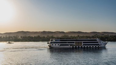Bir turist yolcu gemisi Nil 'de yelken açar. Kıç tarafının arkasında köpük izi var. Sudaki dalgalanmalar. Mavi gökyüzüne karşı kıyı kum tepeleri. Mısır.