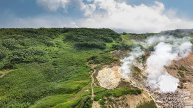 Kaplıcalardan ve fümerollerden gelen buhar dağ yamaçlarından yükseliyor. Yol, yeşil bitki örtüsünün arasındaki tepeye doğru esiyor. Mavi gökyüzündeki bulutlar. Kamçatka.