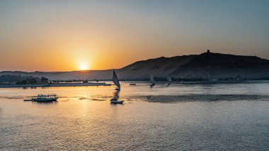 Nil 'de günbatımı. Güneş kum tepelerinin üzerinde batıyor. Gökyüzü turuncuyla işaretlenmiş. Suda güneşli bir patika. Felucca yelkenleri ve tekneler nehirde görülebilir. Mısır